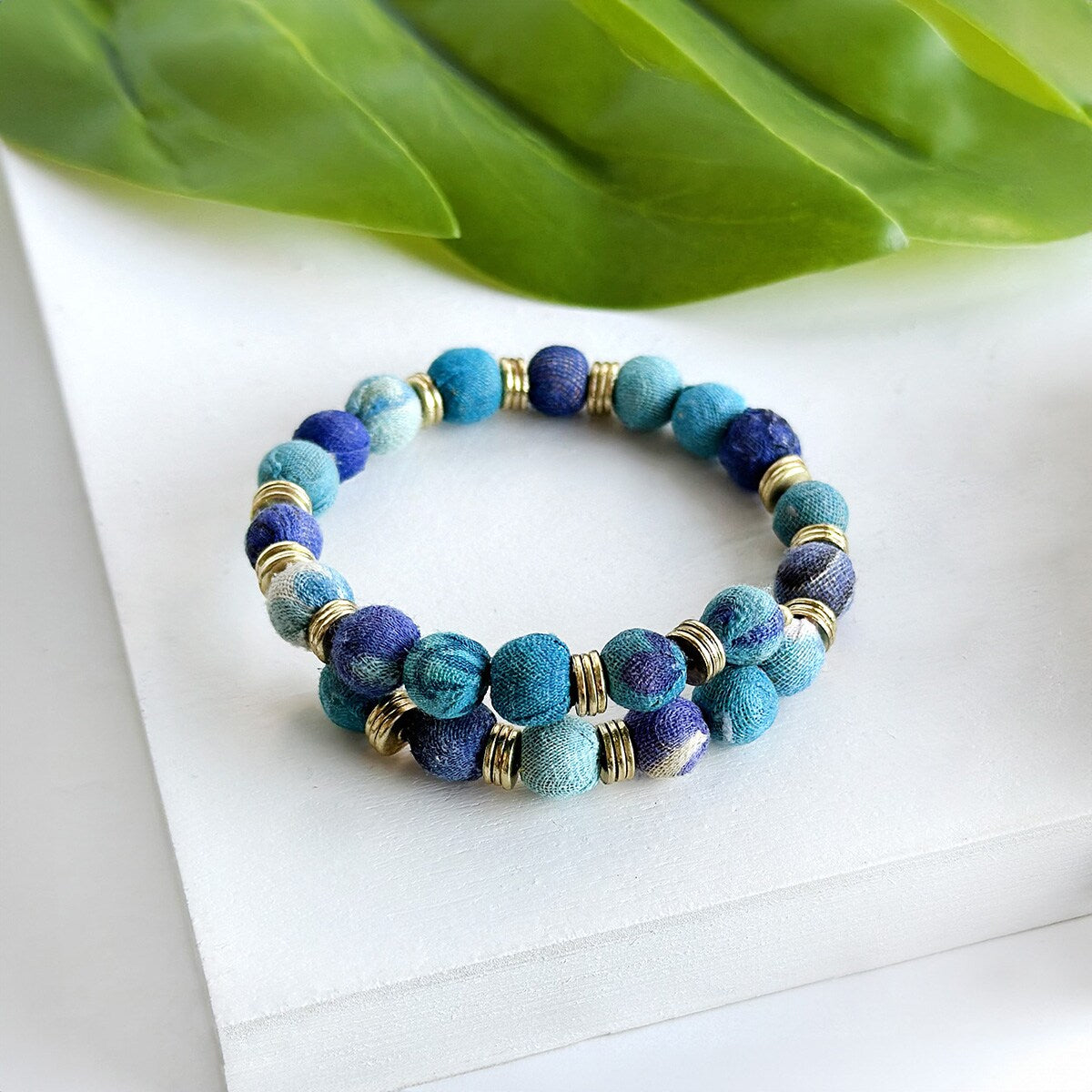 Snap kantha bangle - azure - on white bench with green leaves
