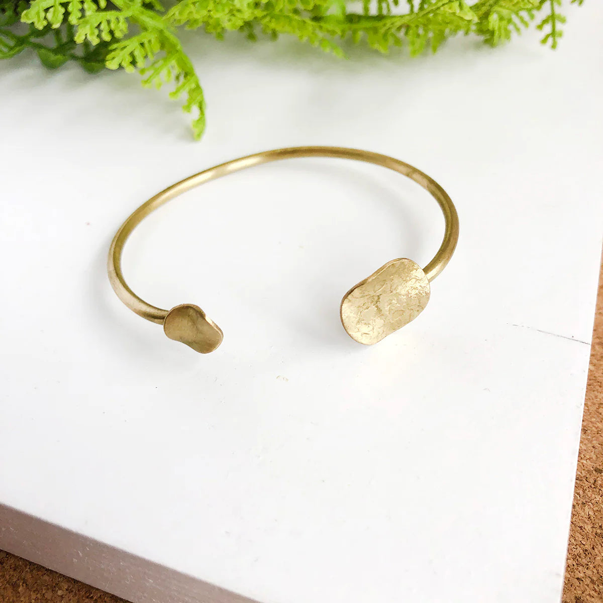 Angelco Accessories hammered coin cuff - gold - flatlay on white board with green fern